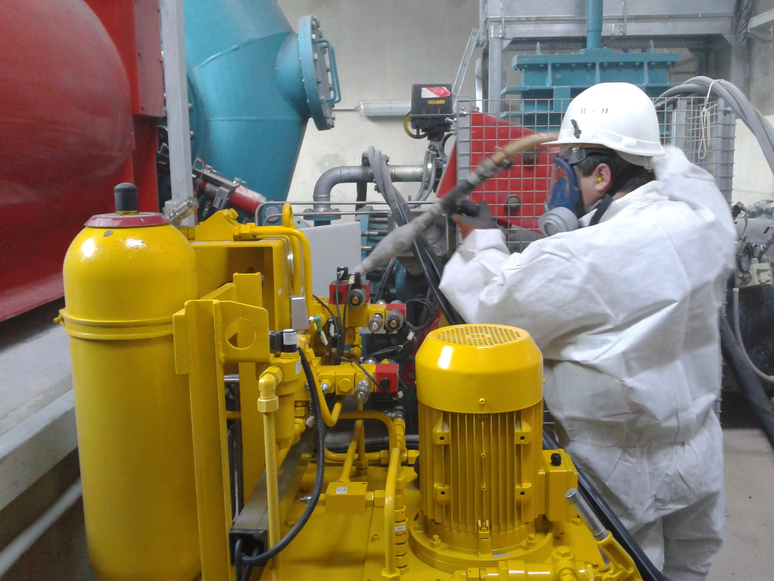 Worker in white protective suit and hard hat operating in industrial cleaning equipment with hydraulic machine at a factory. Technician in protective suit operating cryogenic blasting equipment in a factory environment with yellow machinery and hoses