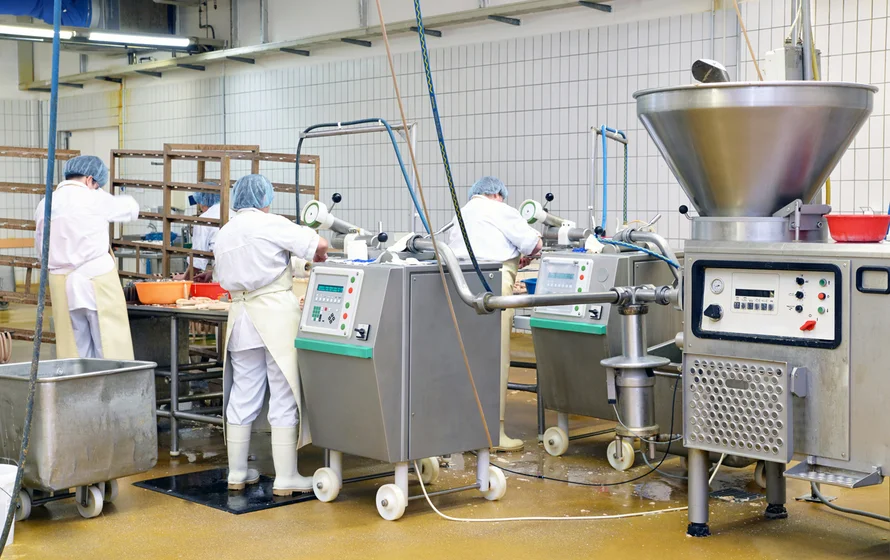 Workers in white coats and hairnets operate ice blasting machinery in a food processing facility, with a large stainless steel hopper and control panels. dry ice blasting food industry